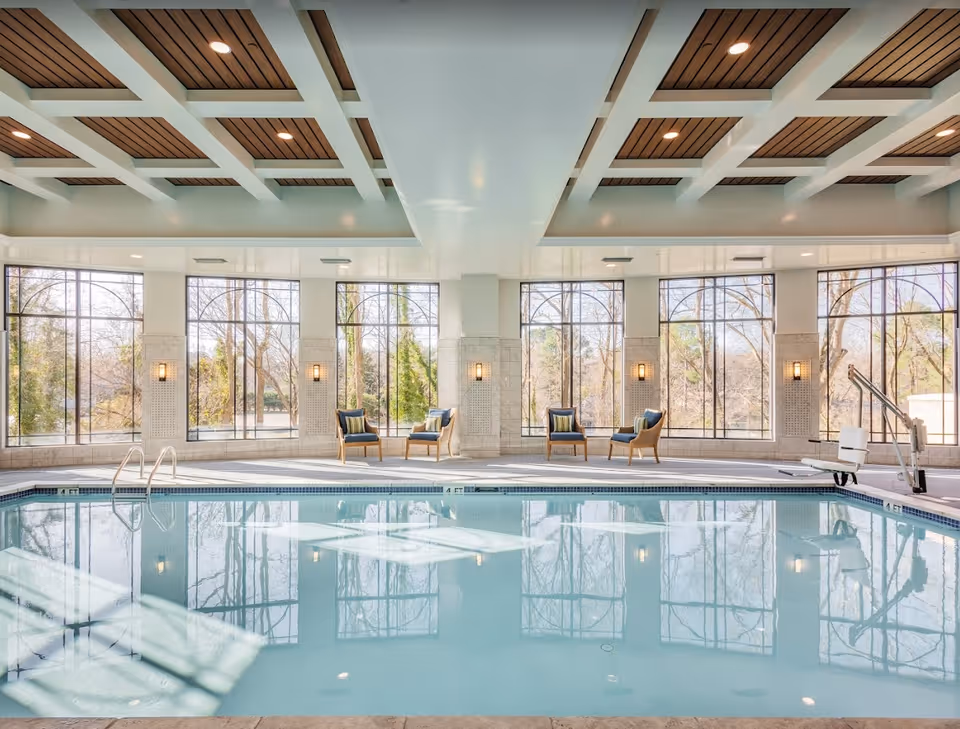 Bright indoor swimming pool with lounge chairs along large arched windows and ceiling accents reflected in the water.