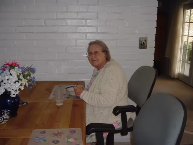 An elderly woman with glasses sits at a wooden dining table eating from a bowl, with a vase of flowers and placemats visible and a white brick wall behind her.