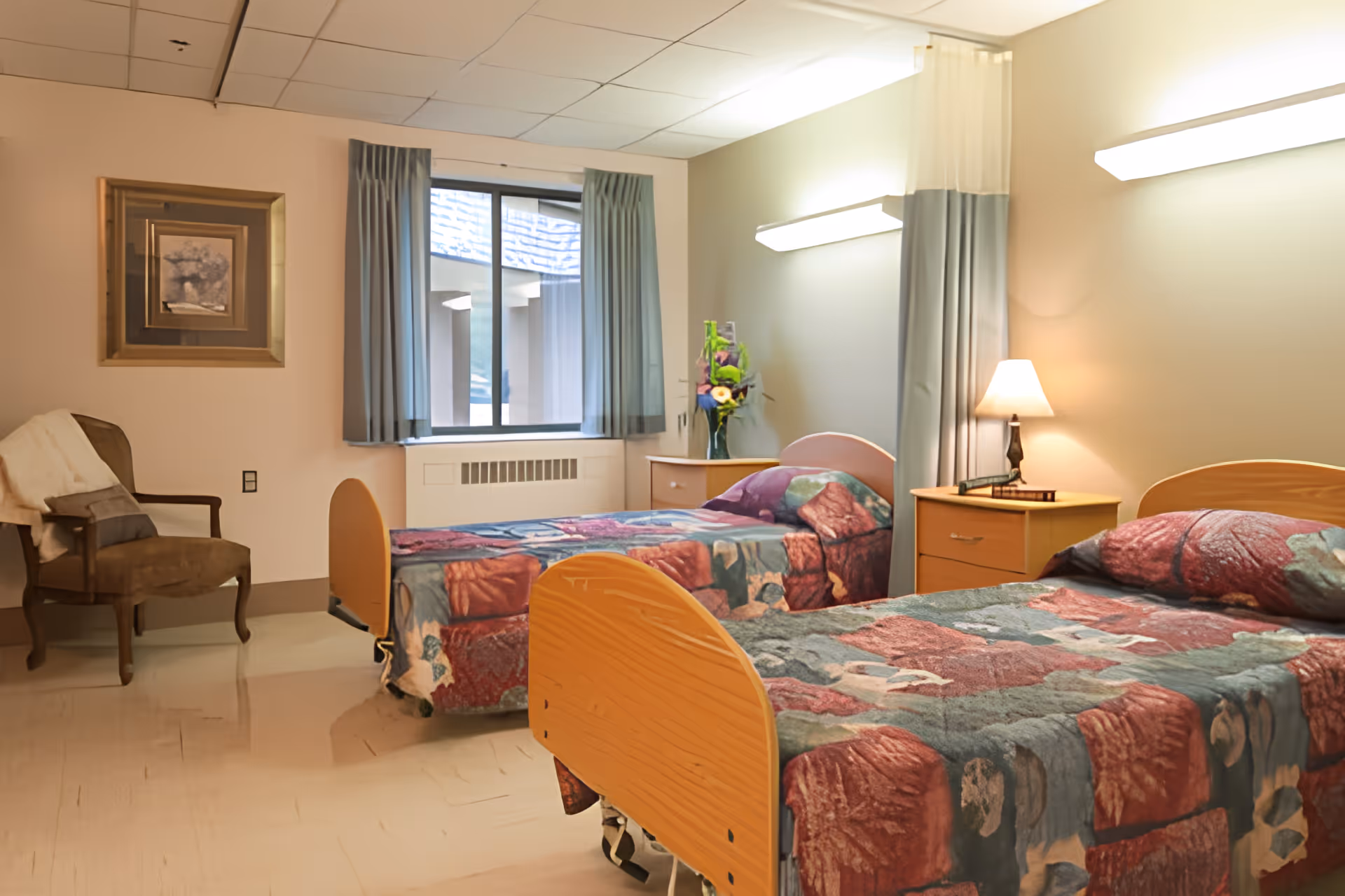 A senior living facility bedroom with two single beds covered in patterned quilts. There is a wooden nightstand with a lamp and a vase of flowers between the beds. A window with blue curtains is on the back wall, and a framed picture hangs on the left wall above a brown armchair with a white throw blanket.