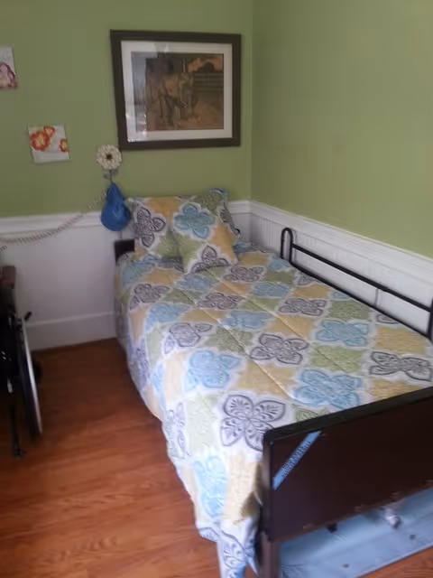 A single bed with a colorful floral patterned bedspread and matching pillow in a small room with light green walls and white wainscoting. There is a framed picture of a horse on the wall above the bed and a blue wall-mounted lamp with a pull cord beside the bed. The floor is wooden.
