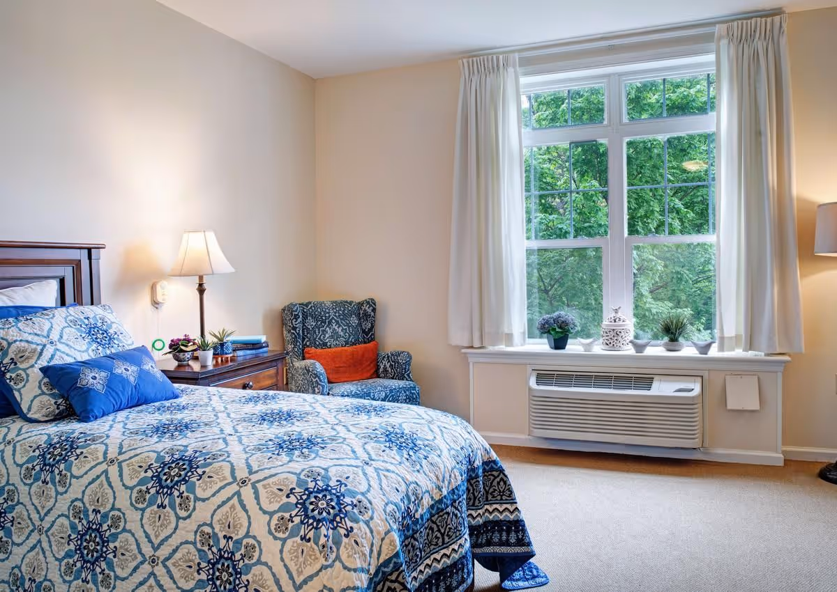 Sunlit bedroom with a patterned blue bedspread, wooden nightstand and lamp, upholstered armchair, and a large window with potted plants above a wall air conditioner.
