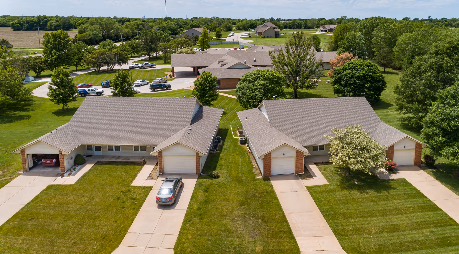 Aerial view of a senior living facility showing two single-story residential buildings with garages, driveways, and well-maintained lawns. In the background, there are additional buildings, parking areas with several cars, and surrounding green trees and open fields under a clear sky.