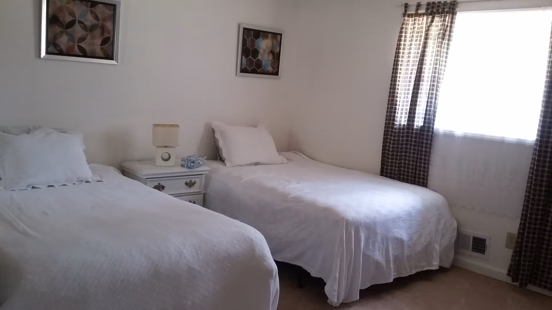 A bedroom with two single beds covered in white bedspreads. Between the beds is a white nightstand with a lamp and a small decorative item. The room has white walls with two framed abstract artworks hanging above the beds. A window with checkered curtains and a sheer white curtain allows natural light into the room.