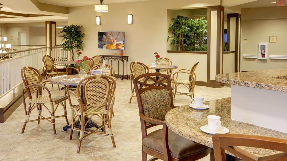 Bright communal dining/lounge area with multiple round tables and wicker chairs beside a granite counter with coffee cups.