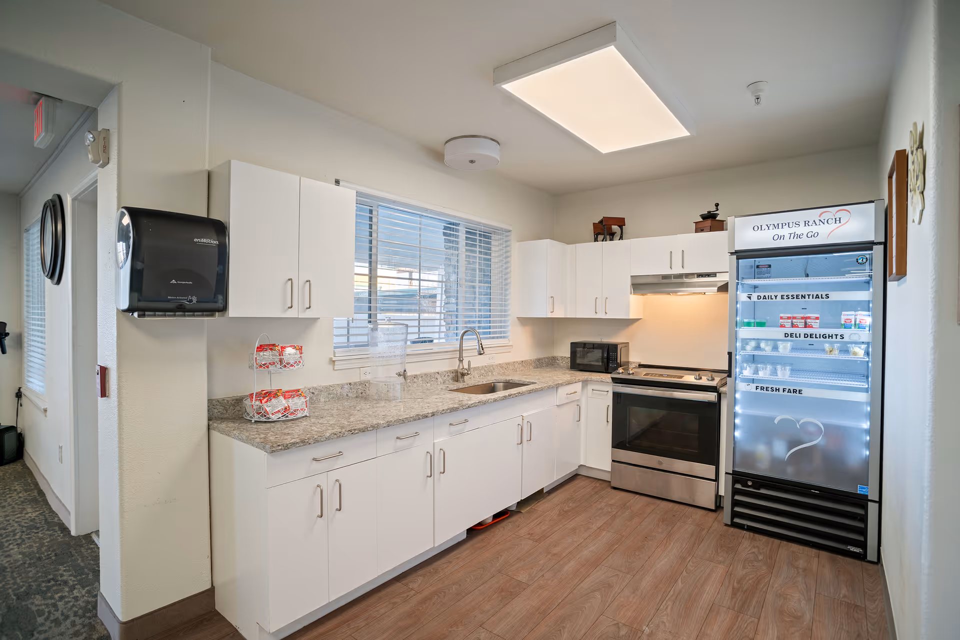 A clean, modern kitchen area with white cabinets, a granite countertop, a stainless steel oven, a microwave, and a refrigerator labeled 'Olympus Ranch On The Go' stocked with various food items. There is a window with blinds above the sink and a paper towel dispenser mounted on the wall.