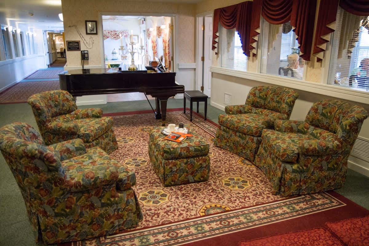 A cozy sitting area with four floral upholstered armchairs arranged around a matching ottoman on a patterned area rug. Behind the seating area is a black grand piano with a candelabra on top. The room has large windows with red valances and a hallway visible in the background.