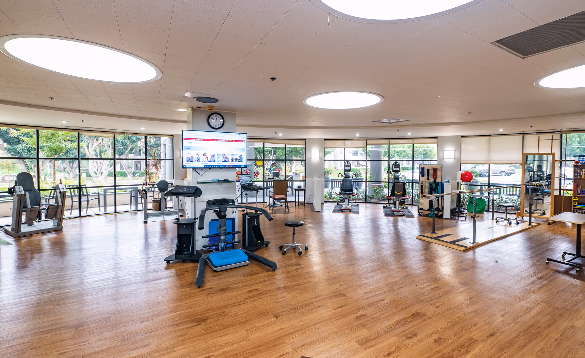Spacious exercise and therapy room in a senior care facility with workout equipment, large windows, and wood flooring.
