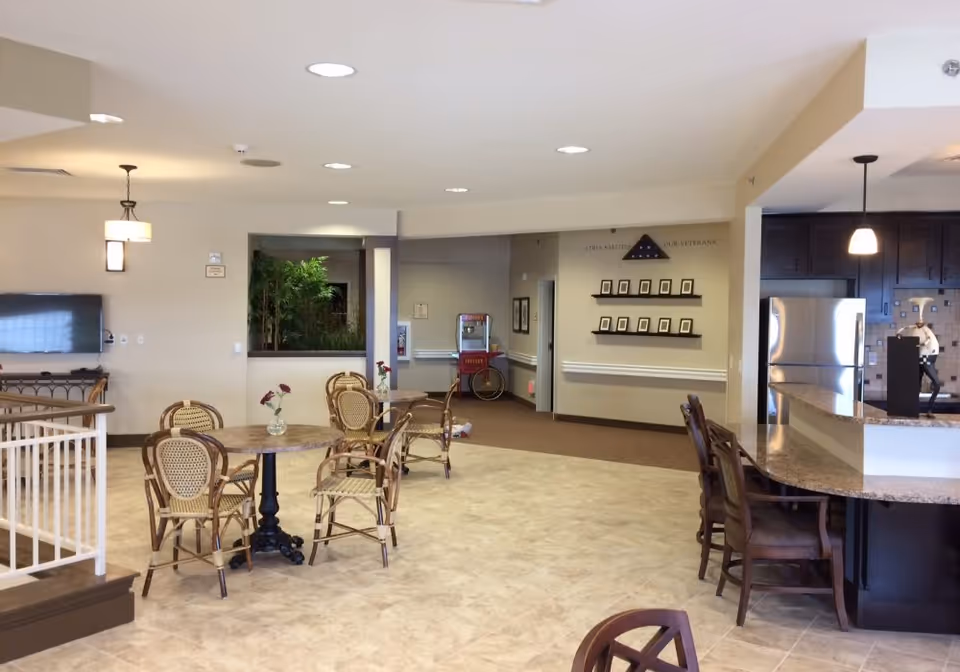 Open senior living common area with round tables and chairs, a counter with bar stools, and a kitchenette in the background.