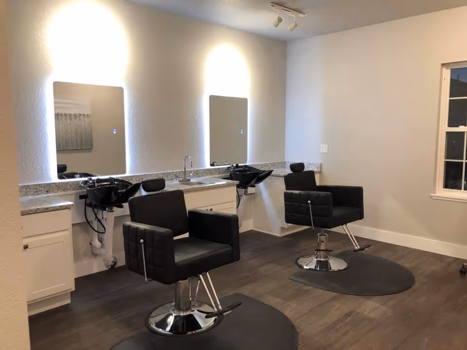 Interior view of a salon area with two black salon chairs in front of two sinks and mirrors mounted on a white wall, with overhead lighting and a window on the right side.