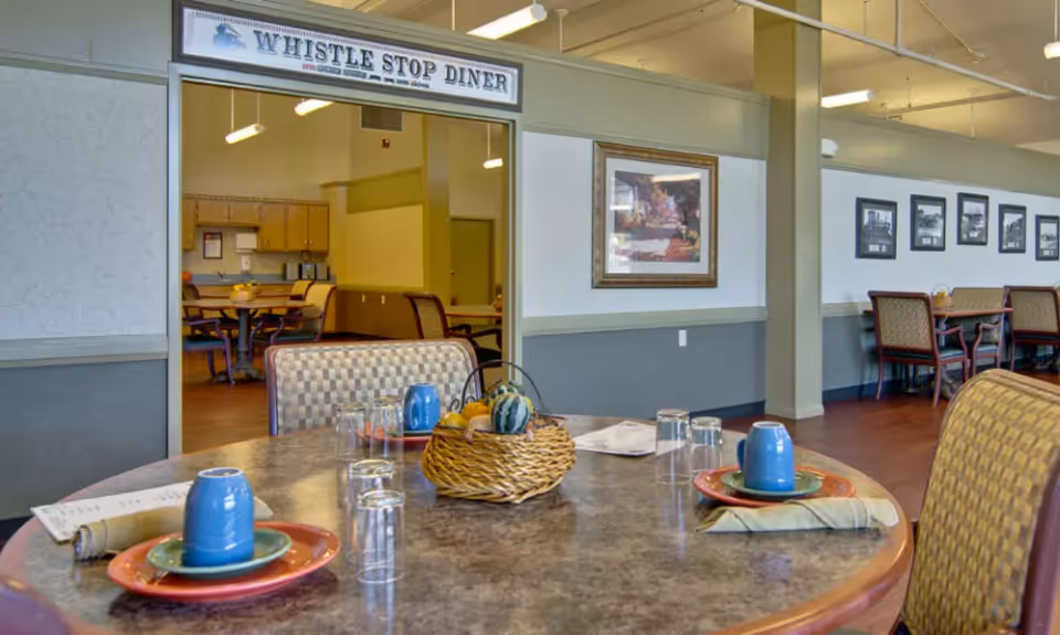 Interior view of a dining area named Whistle Stop Diner with tables set with plates, blue cups, napkins, and glasses. The room has framed pictures on the walls and a basket with decorative gourds on the table in the foreground.