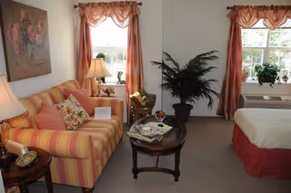 A cozy living area with a striped sofa adorned with floral pillows, a wooden coffee table with magazines and decorative items, a potted plant, and two windows with sheer curtains letting in natural light. A bed with a red bed skirt is partially visible on the right side of the room.
