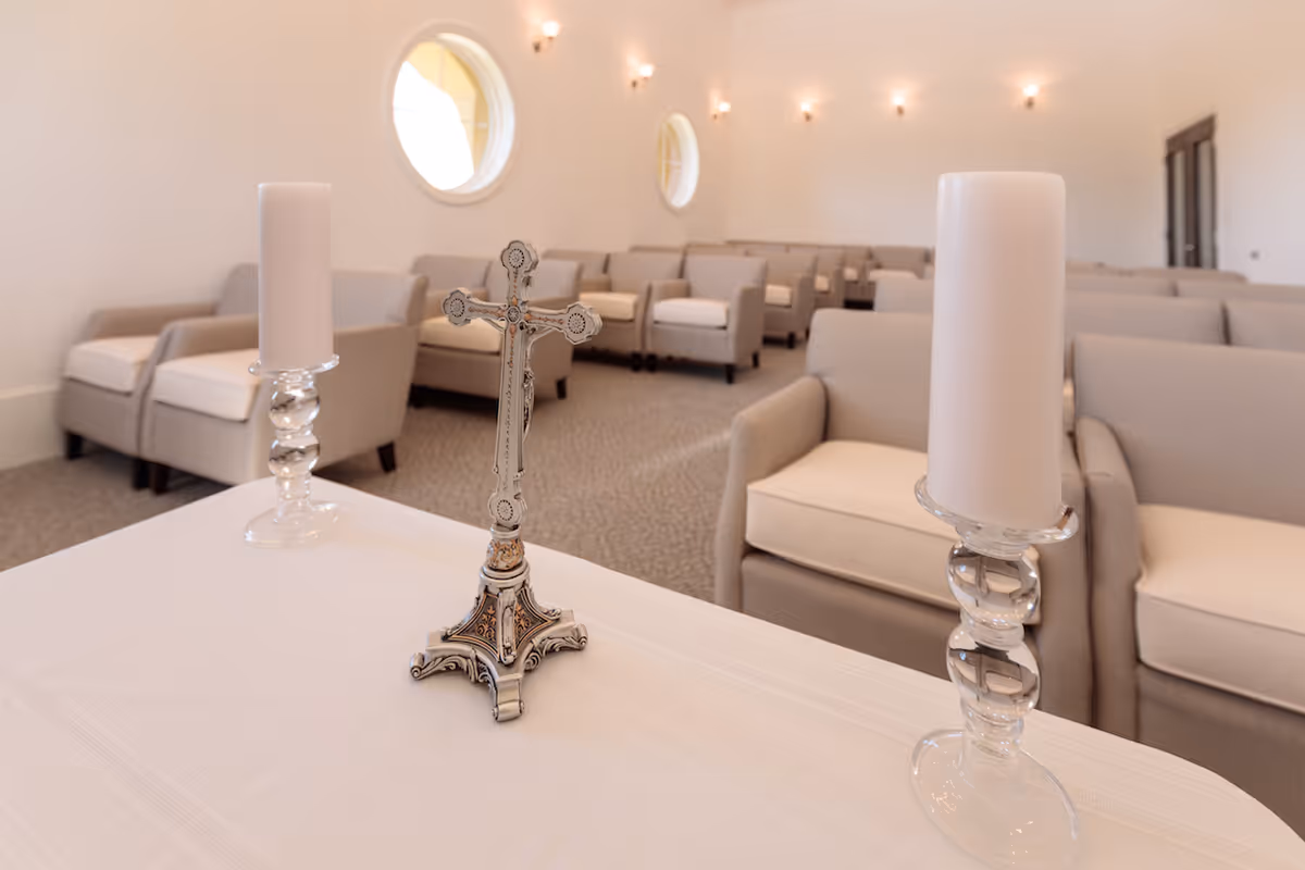 Interior view of a chapel or meditation room with rows of beige armchairs, a white table at the front holding a decorative silver cross and two white candles in glass holders, and round windows on the wall.