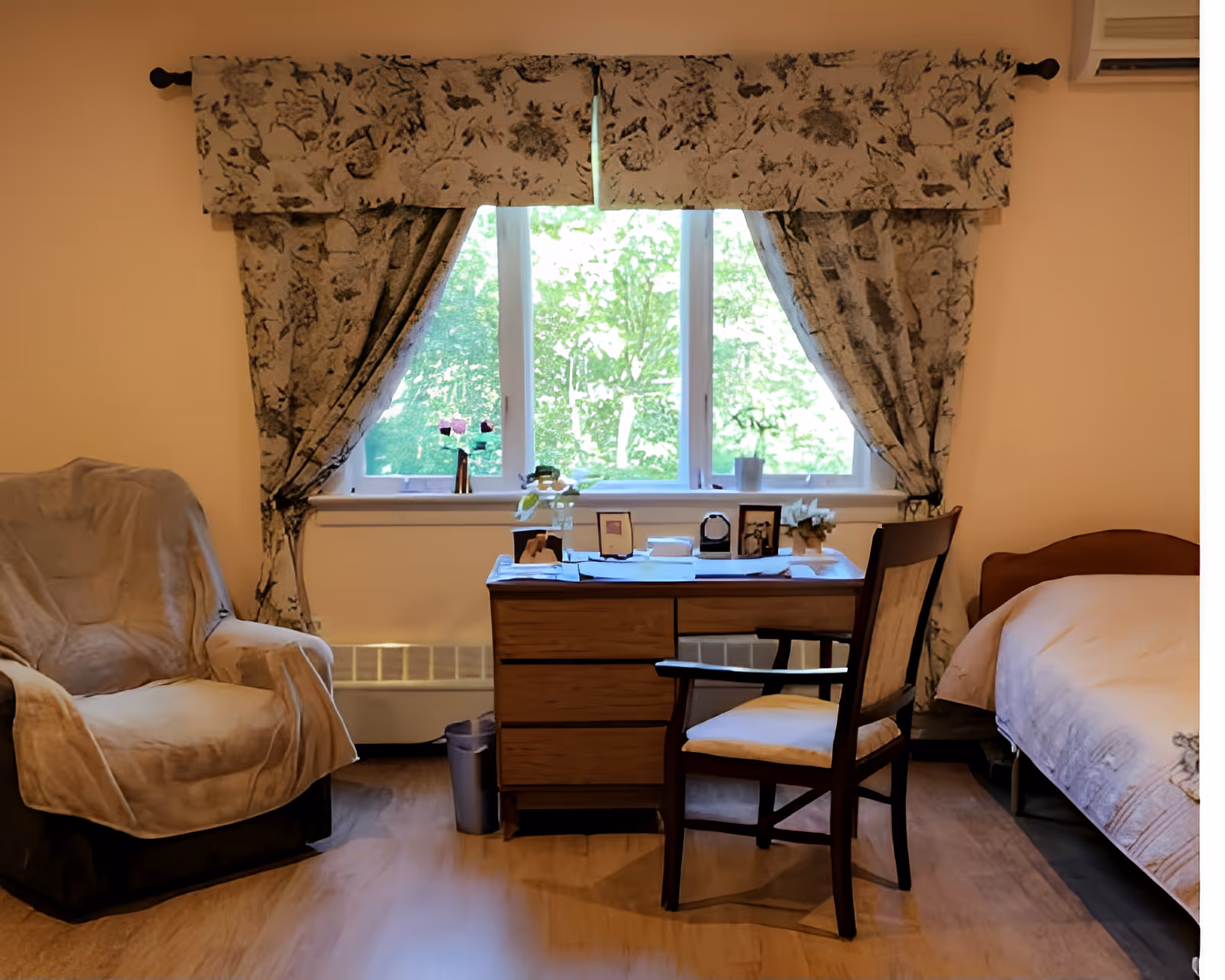 A cozy bedroom with a desk and chair in front of a window flanked by a covered armchair and a bed.