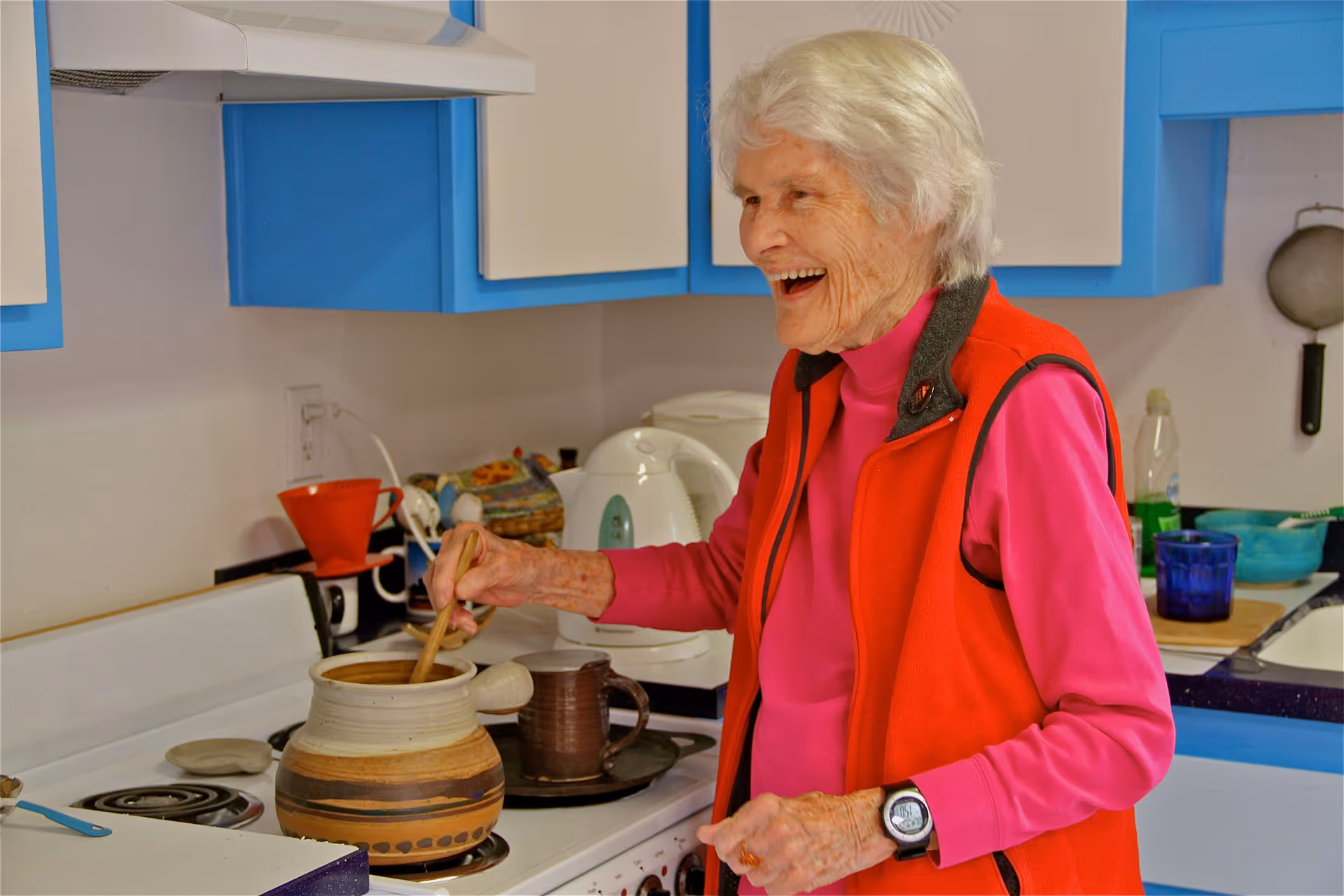 An elderly woman with white hair, wearing a pink shirt and red vest, is smiling and stirring a pot on a stove in a kitchen with blue and white cabinets.