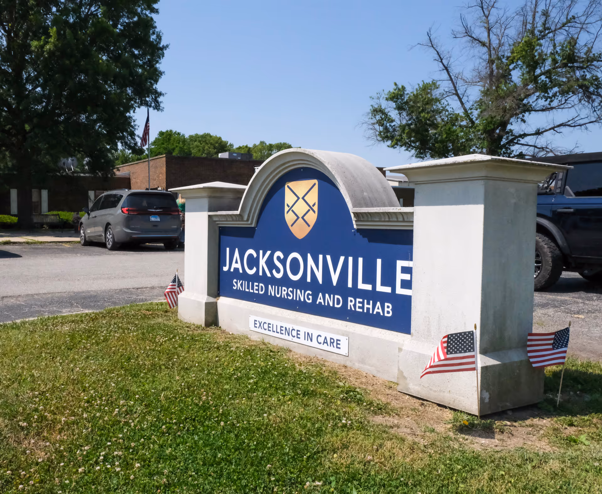 Exterior view of the Jacksonville Skilled Nursing and Rehab entrance sign on a grassy lawn with small American flags, parked cars, and the facility building and trees in the background.