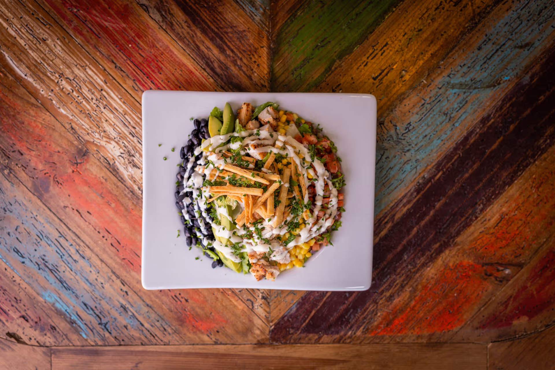 Top view of a square white plate with a colorful salad consisting of black beans, avocado slices, grilled chicken, corn, diced tomatoes, tortilla strips, and drizzled with a creamy dressing, placed on a rustic multicolored wooden table.