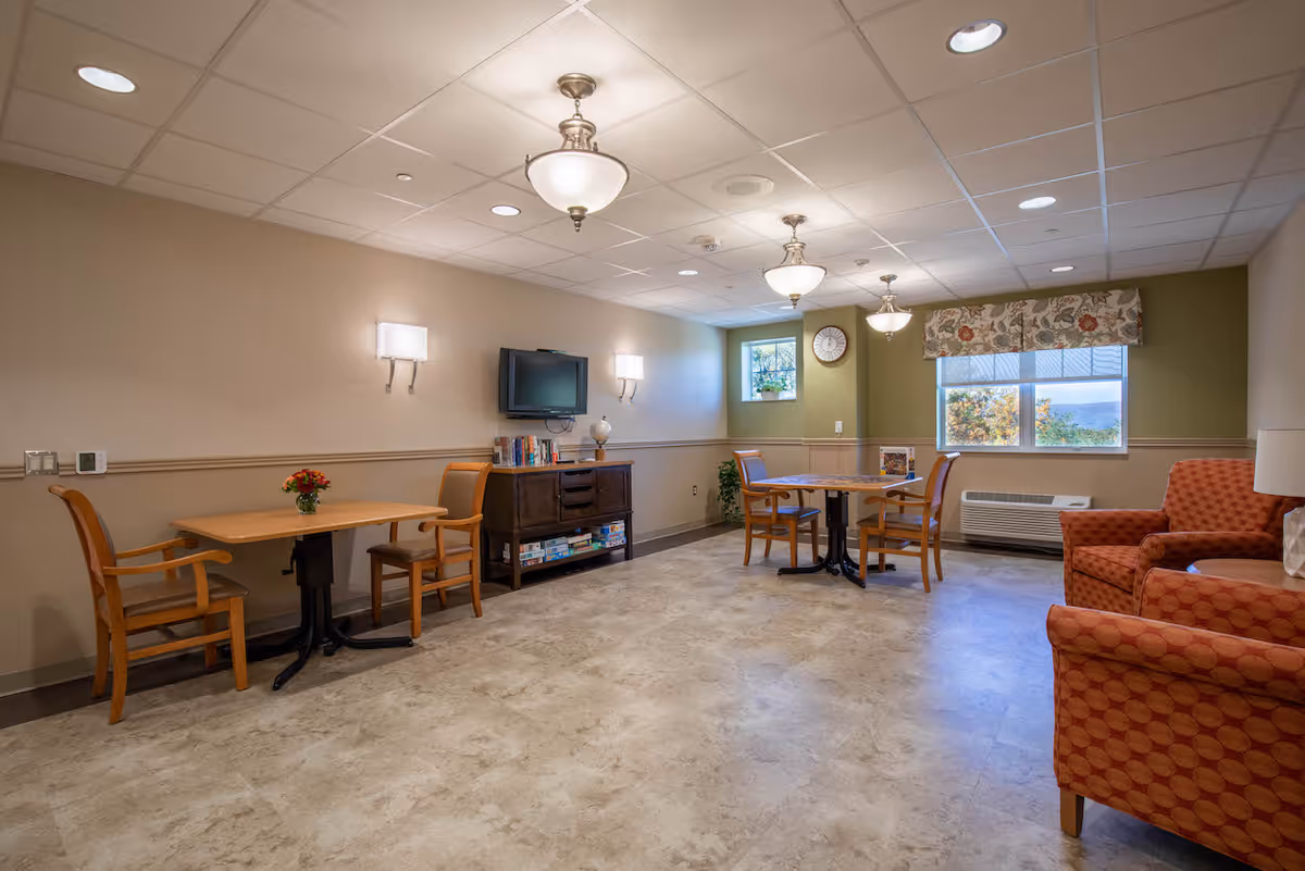 A well-lit common room in a nursing and rehabilitation center featuring two small wooden tables each with two chairs, a wall-mounted TV above a cabinet with books and board games, two patterned armchairs, a window with floral valance, and ceiling light fixtures.