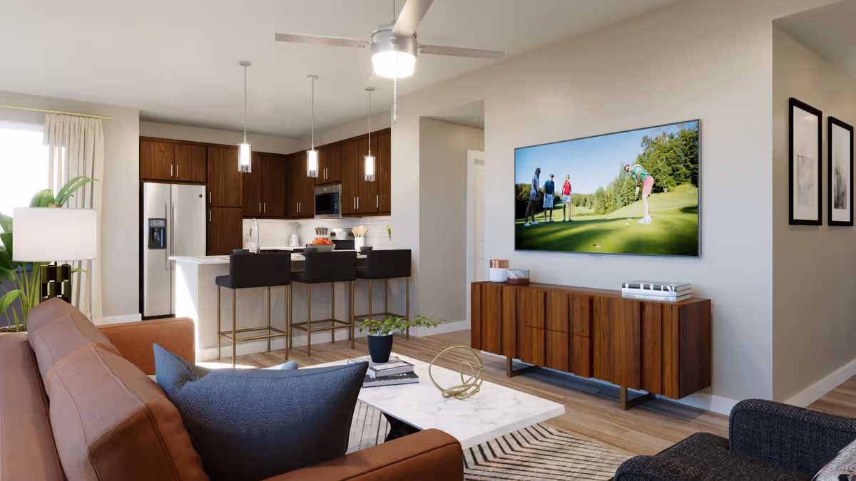 A modern open-concept living room and kitchen area featuring a brown sofa with blue pillows, a white marble coffee table with decorative items, a wooden media console with a flat-screen TV mounted above it displaying a golf scene, and a kitchen with dark wood cabinets, stainless steel refrigerator, and three black bar stools at the kitchen island. The room is well-lit with natural light from a window with curtains and pendant lights above the kitchen island.