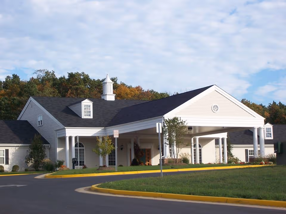 Exterior view of a single-story building with white siding and a black roof, featuring a covered entrance supported by white columns. The building is surrounded by a paved driveway, green grass, and trees with autumn foliage in the background under a partly cloudy sky.