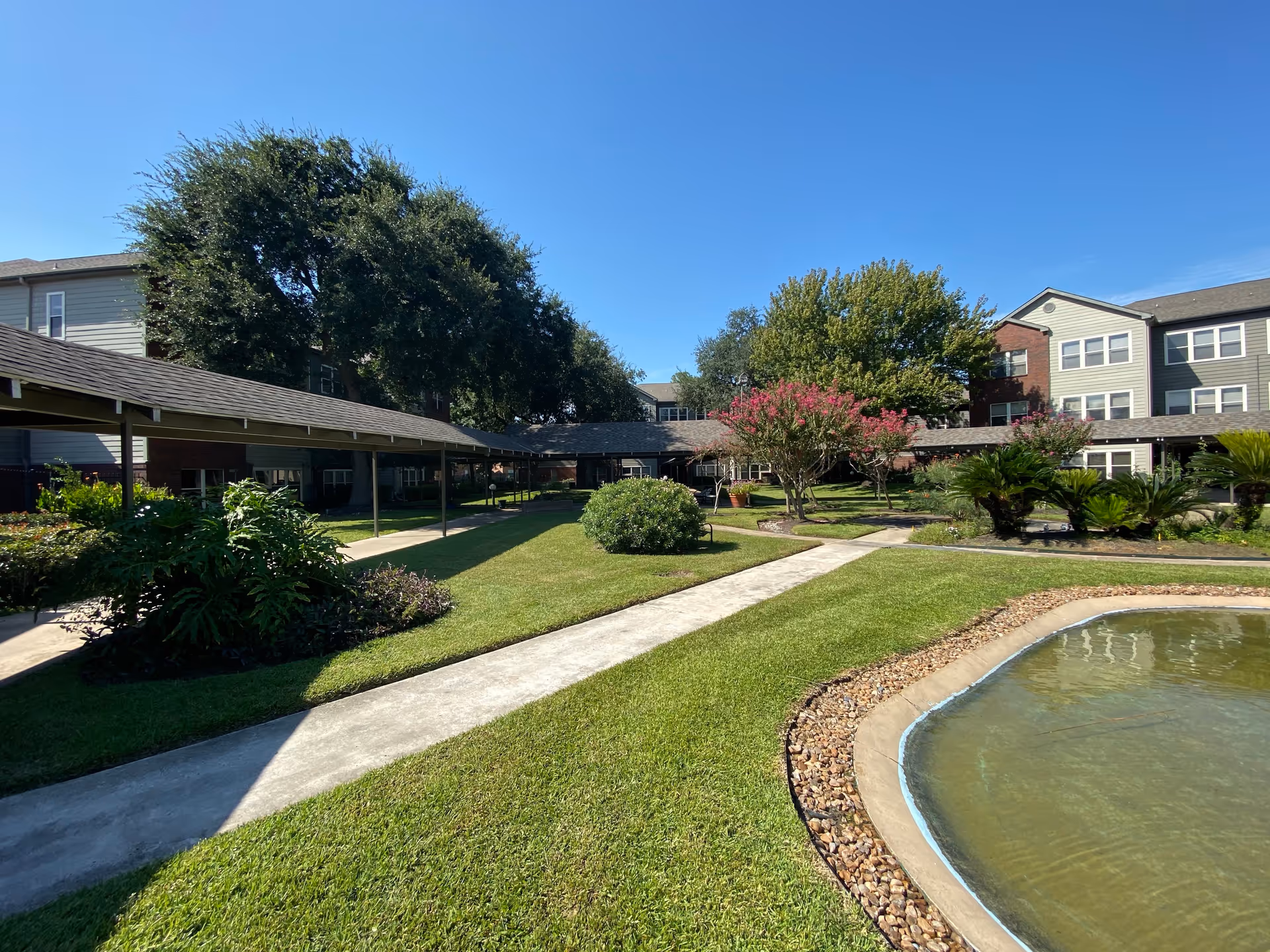 Outdoor courtyard area of a senior living facility with well-maintained green lawns, a small pond edged with stones, various trees and shrubs including flowering plants, and covered walkways connecting multi-story residential buildings under a clear blue sky.