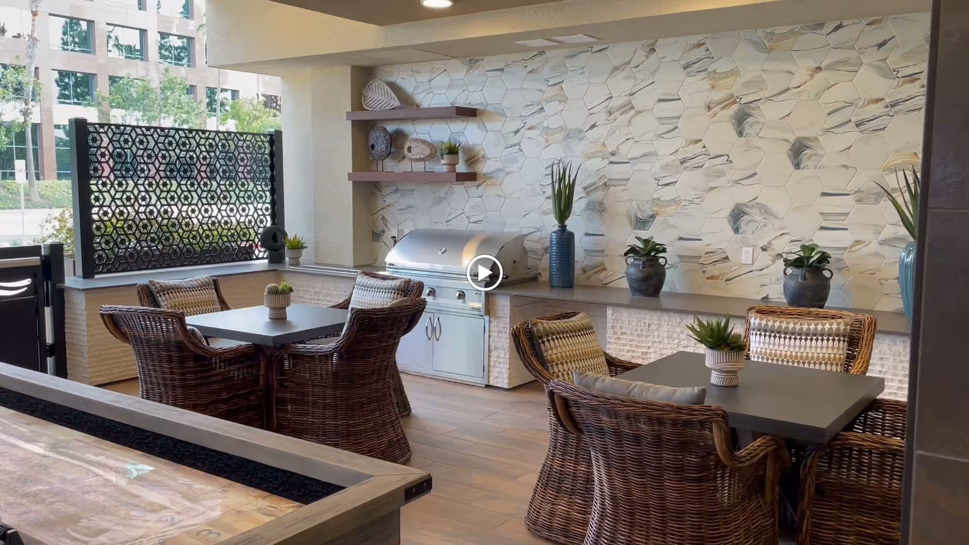 Outdoor seating area with two tables surrounded by wicker chairs, decorative plants on the tables and shelves, a built-in grill, and a patterned tile wall in the background.