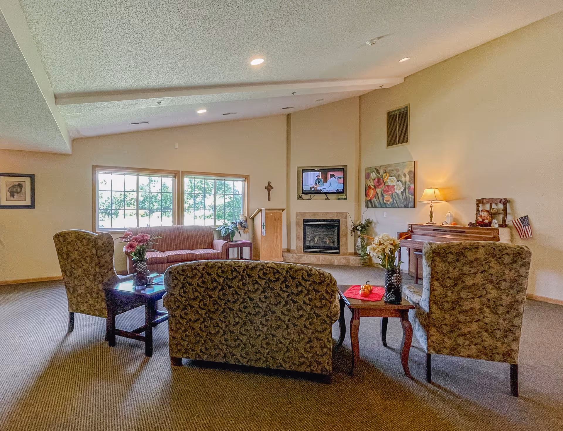 A cozy living room area with patterned armchairs and a sofa arranged around a coffee table with flowers. There is a fireplace with a mounted TV above it, a piano with a lamp and decorations, and large windows letting in natural light.