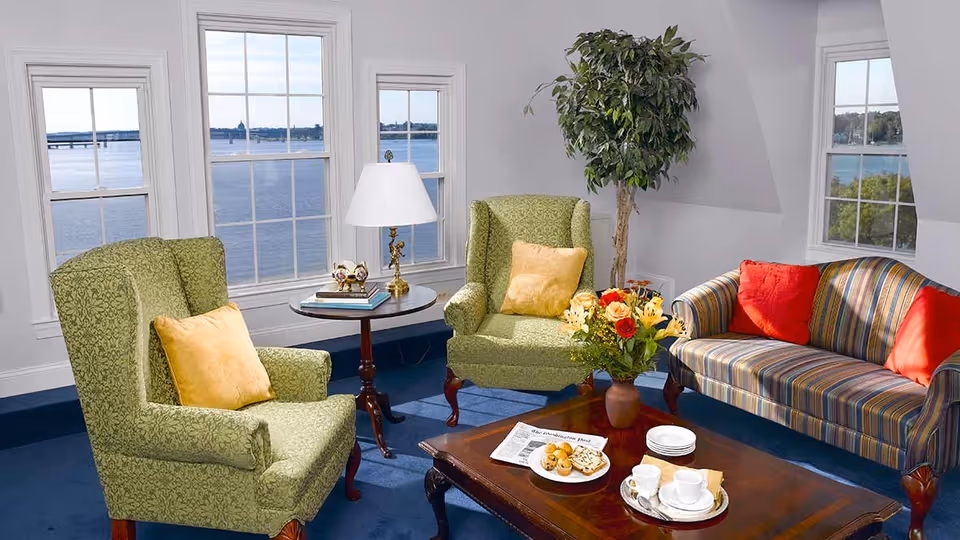 A cozy living room with two green patterned armchairs each with a yellow pillow, a striped sofa with red pillows, a wooden coffee table with a vase of flowers, a plate of snacks, and a tea set. There is a side table with a lamp and decorative items between the armchairs. Large windows show a scenic view of a body of water and a bridge outside.