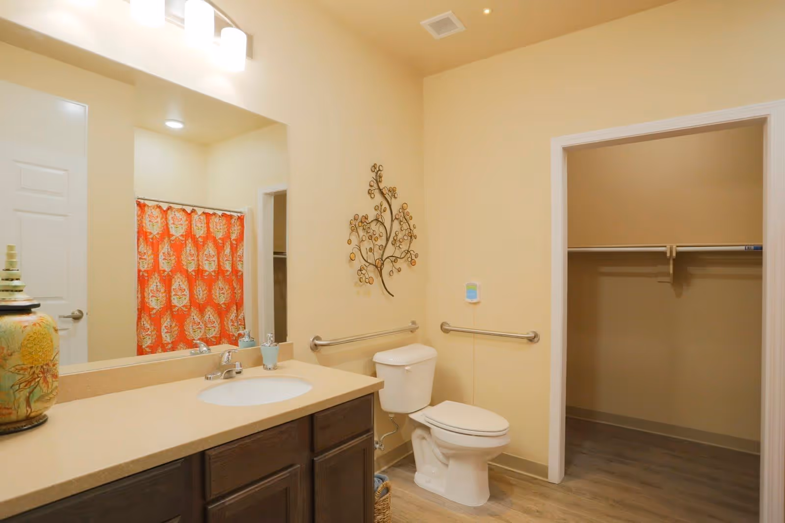 A bathroom with a beige countertop and sink, a large mirror above the sink, a white toilet with grab bars on the walls beside it, a decorative metal wall art above the toilet, a walk-in closet with wooden flooring, and a shower with a red and orange patterned curtain.