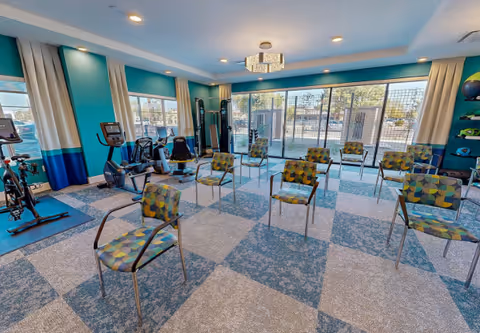 A bright exercise room with patterned chairs arranged in rows on a checkered carpet floor. The room has large windows and glass doors letting in natural light, with exercise equipment such as stationary bikes and weight machines along the walls. The walls are painted teal with white trim, and there is a modern ceiling light fixture.