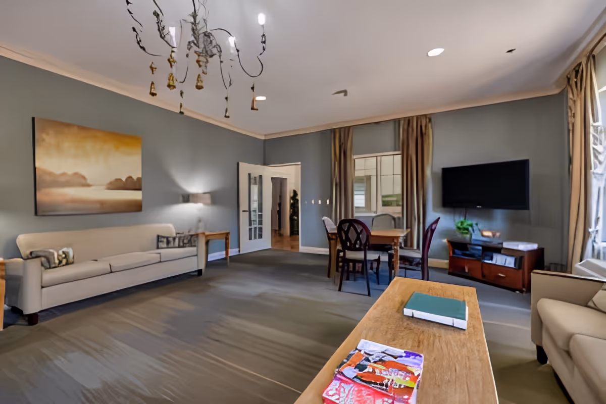 A spacious living room with light gray walls and carpeted floor. The room features a beige sofa along the left wall, a wooden coffee table with books on it in the foreground, a dining table with chairs near the back wall, a flat-screen TV mounted on the right wall above a wooden TV stand, and large windows with curtains allowing natural light to enter. A large painting hangs above the sofa, and a chandelier is visible on the ceiling.