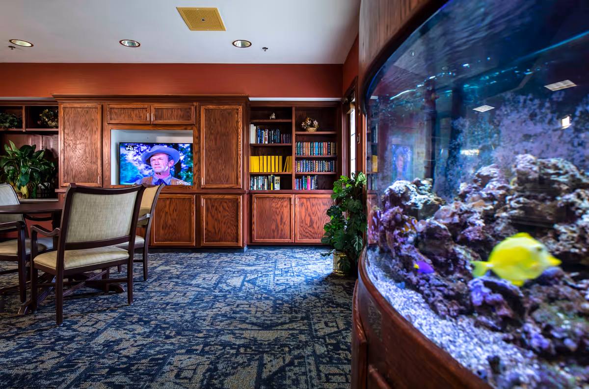 Interior view of a common area in an assisted living facility featuring a large curved aquarium with colorful fish on the right, a carpeted floor with a blue patterned design, wooden bookshelves filled with books and decorative items, a television displaying a man wearing a hat, and a table with chairs in the foreground.