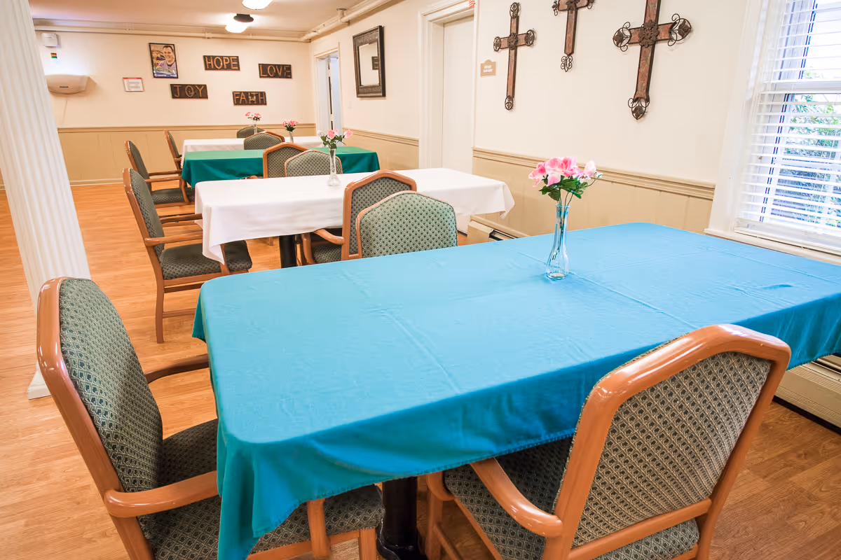 A dining room with several tables covered with blue and white tablecloths, each table having a small vase with pink flowers. The room has wooden flooring, green cushioned chairs with wooden frames, and walls decorated with crosses and wooden plaques with words like HOPE, LOVE, JOY, and FAITH. A window with blinds allows natural light into the room.