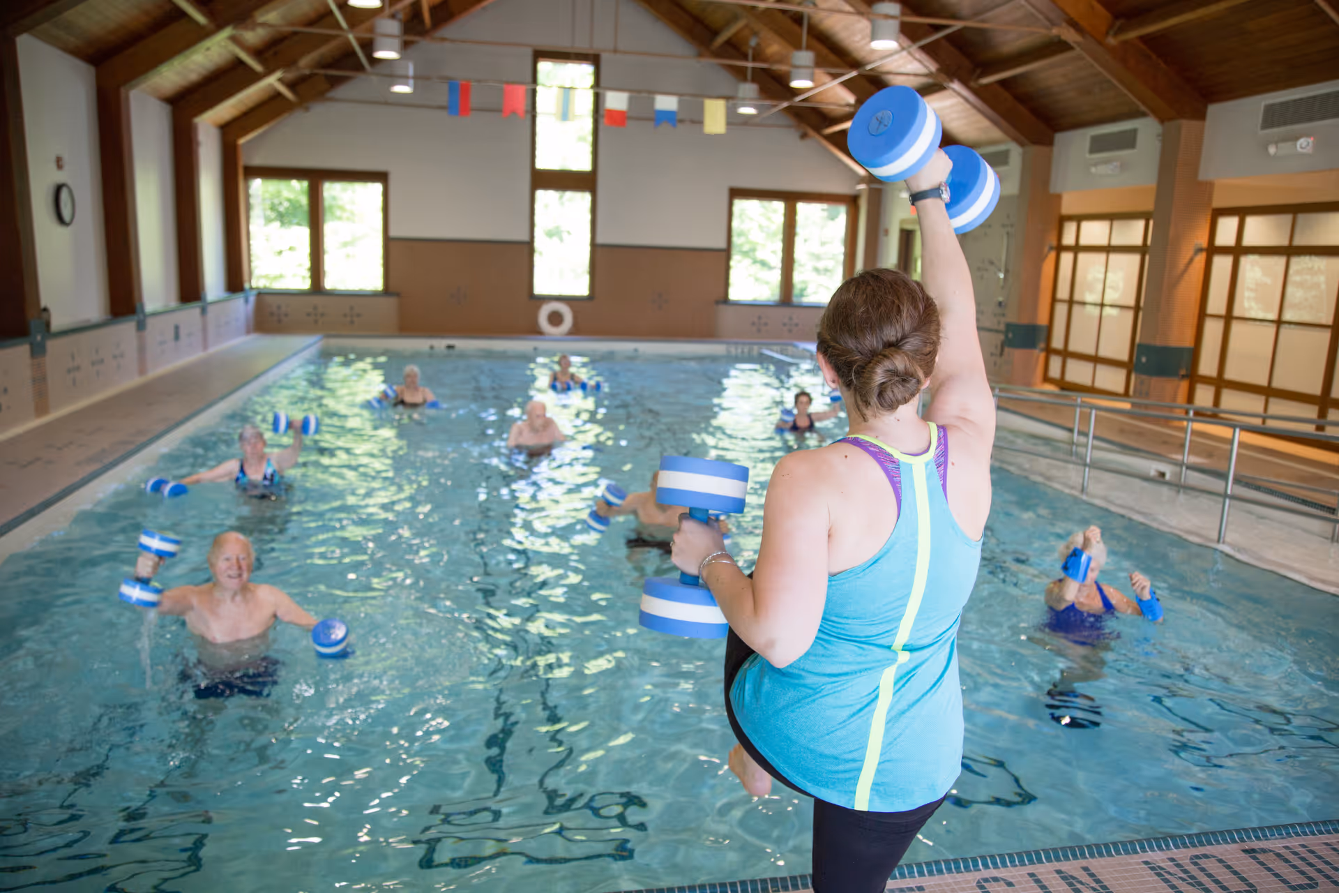 An indoor swimming pool with a group of elderly people participating in a water exercise class led by an instructor holding blue and white foam dumbbells. The pool area has large windows and a wooden ceiling with exposed beams.