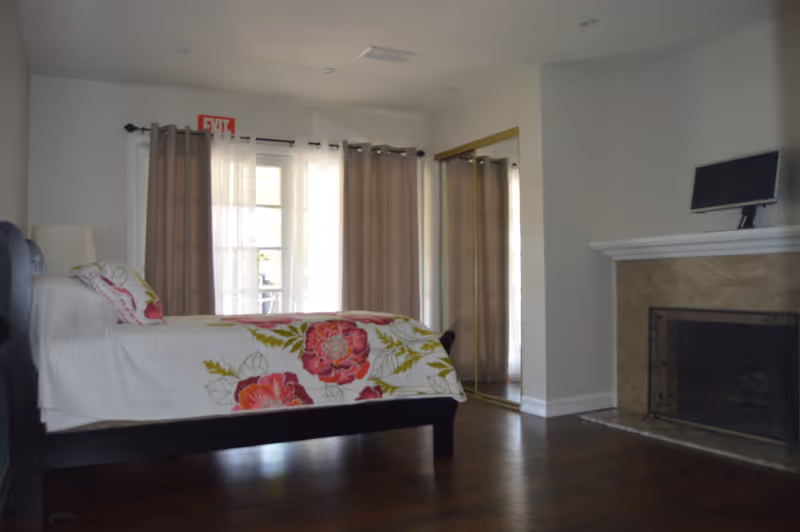 A bedroom with a dark wooden bed frame and a white bedspread featuring large pink and red floral patterns. The room has hardwood floors, a fireplace with a beige stone surround, and a small TV on the mantel. There are large windows covered with sheer white and beige curtains, and a mirrored closet door on the right side of the room.