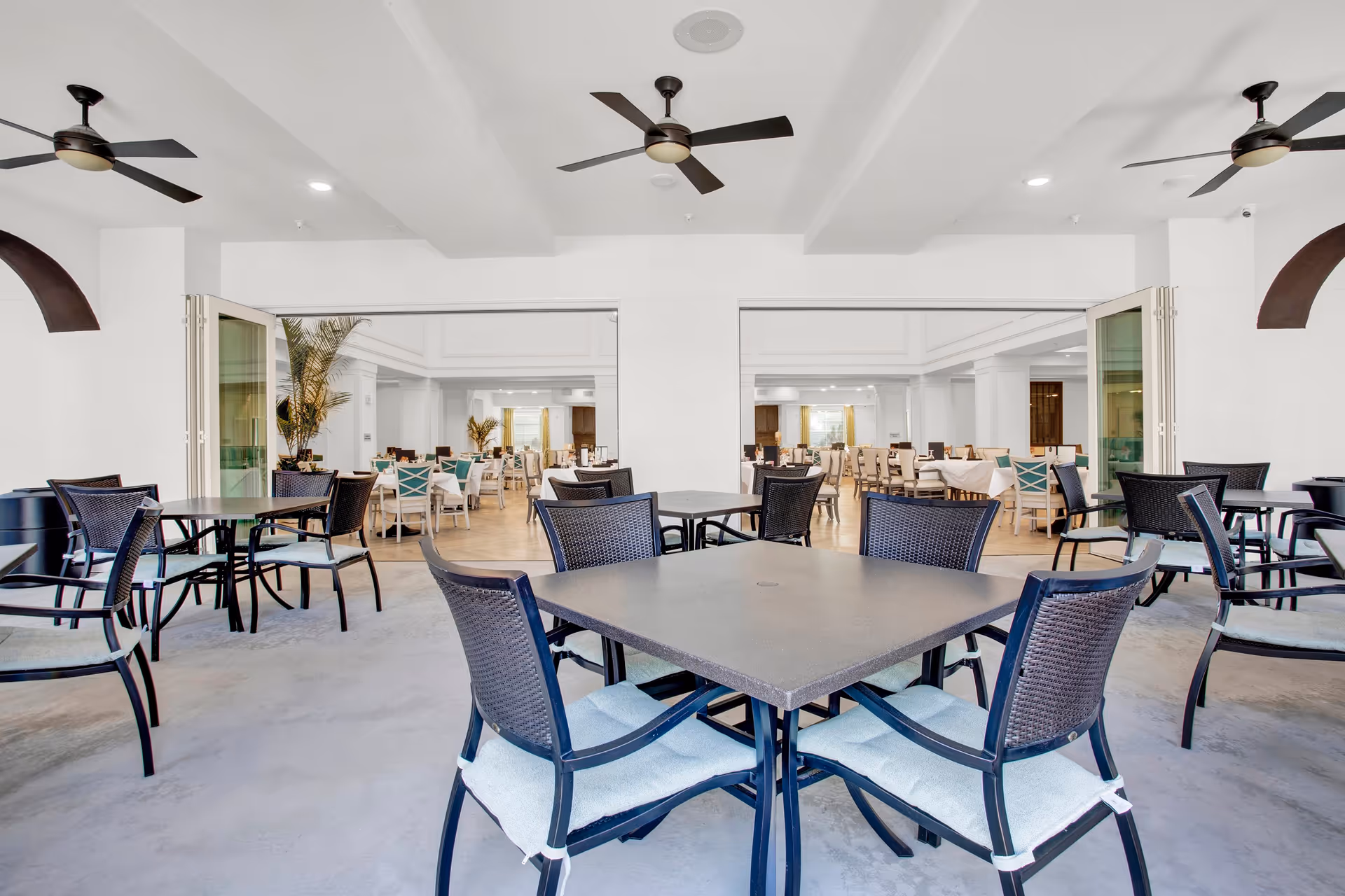 A spacious dining area with multiple tables and chairs. The foreground shows a patio-style seating area with dark metal tables and chairs with light cushions. The background reveals an indoor dining room with white walls, ceiling fans, and additional tables and chairs arranged neatly. Large open doorways connect the indoor and outdoor spaces, and there are potted plants adding greenery to the setting.