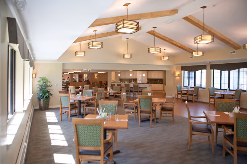 A spacious dining room in a senior living facility with multiple wooden tables and chairs arranged neatly. The room features large windows on the left side allowing natural light to fill the space, ceiling lights with wooden beams, and a kitchen area visible in the background. There is a potted plant near the windows and the overall decor is warm and inviting.
