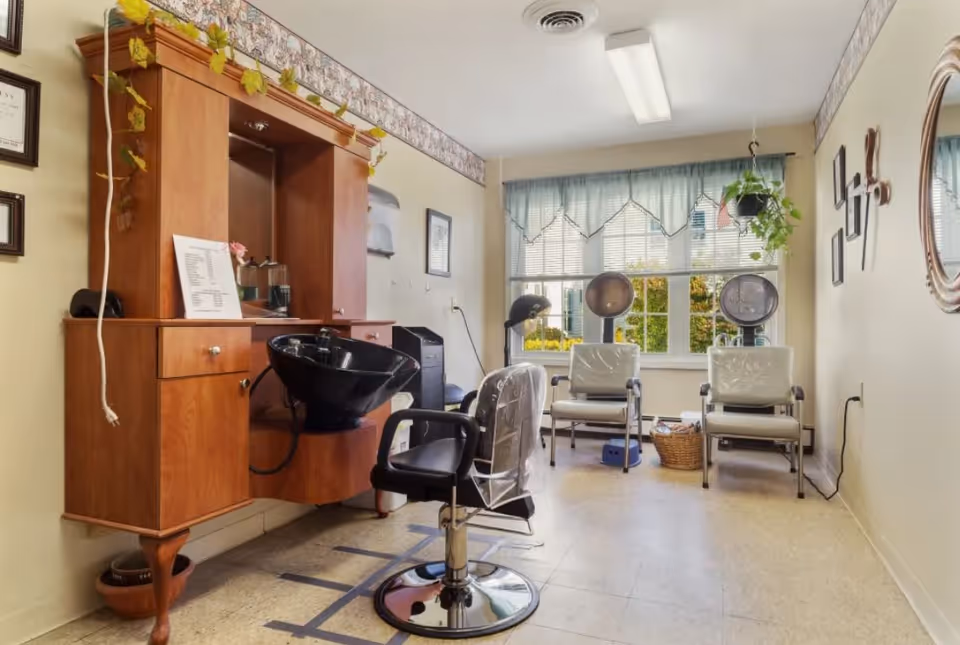 A small hair salon area inside a senior living facility with a wooden cabinet holding a black wash basin, a salon chair in front, and two white chairs with hair drying hoods near a window with blue curtains. The room has light-colored walls with framed pictures and a round mirror on one side, and a hanging plant near the window.