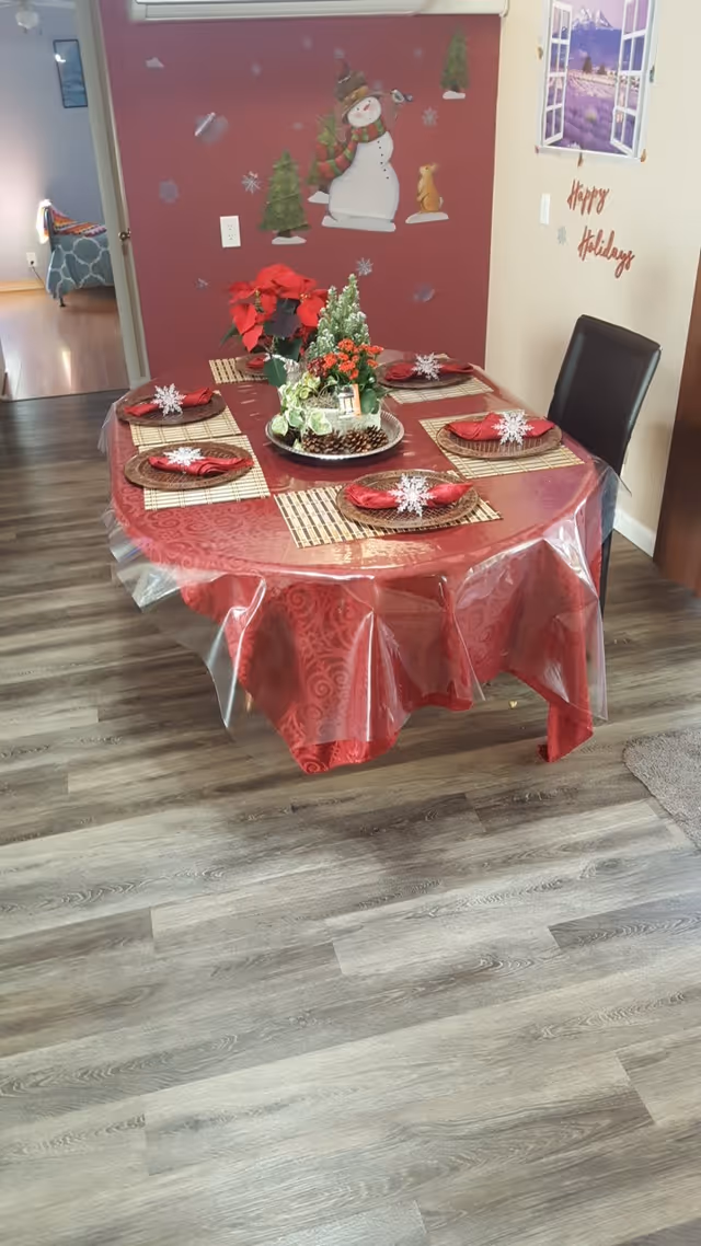 Round dining table set for six with a red tablecloth and holiday centerpiece in a dining area with wood-look flooring and festive wall decals.
