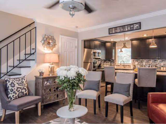 A cozy living area with a small round table holding a vase of white flowers, surrounded by four upholstered chairs. To the left is a wooden side table with a lamp and decorative items, next to a staircase with black railings. In the background is a modern kitchen with dark cabinets, stainless steel appliances, a tiled backsplash, and a breakfast bar with three high chairs.
