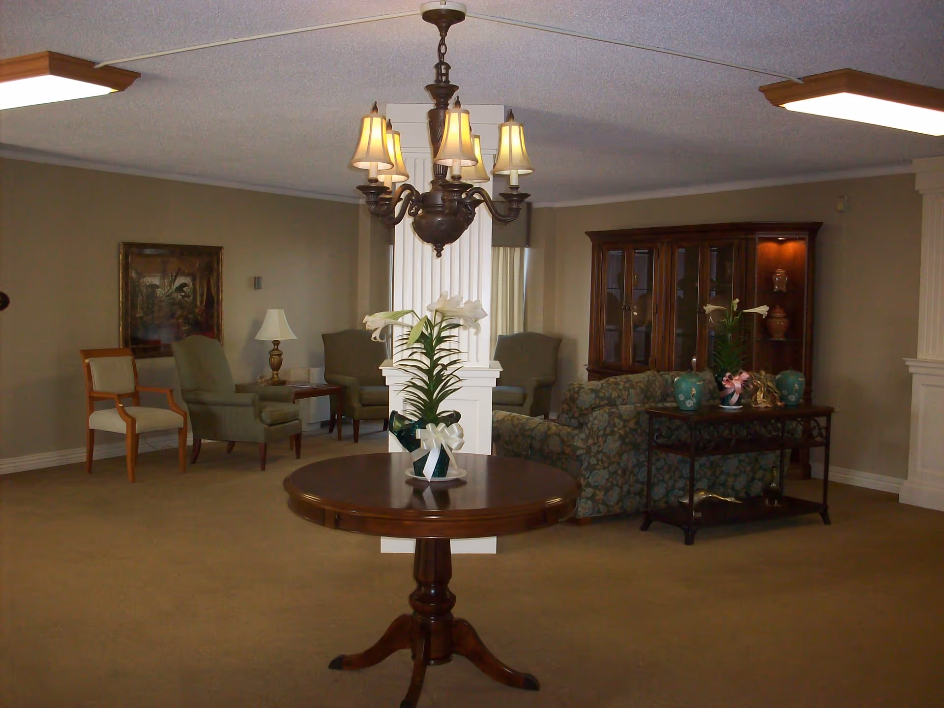 A senior living facility common area with beige carpet and walls. In the center is a round wooden table with a white flower arrangement. Behind it is a white column and a chandelier with six lampshades. To the right is a floral patterned sofa and a wooden display cabinet with glass doors. To the left are several upholstered chairs and a side table with a lamp. The room is softly lit with ceiling lights.