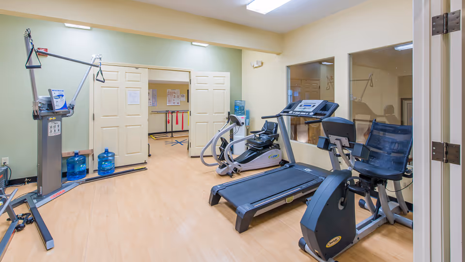 Fitness room with a treadmill, stationary/recumbent bikes, resistance machine, and other exercise equipment on a light wood floor.
