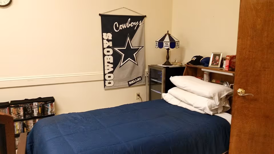 A small bedroom with a single bed covered in a blue blanket and stacked white pillows. On the wall above the bed hangs a Dallas Cowboys banner. Next to the bed is a small nightstand with a lamp, a framed photo, a hat, and some other personal items. A wooden door is partially open on the right side of the image.