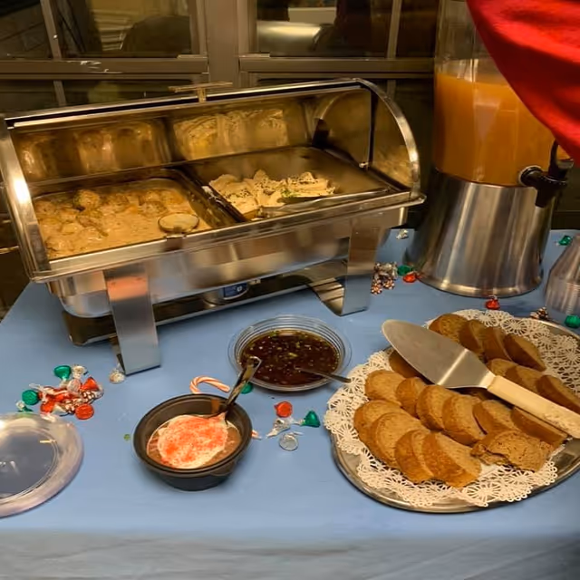 Buffet table with a stainless steel chafing dish containing two trays of food, a bowl of sauce with a spoon, a plate of sliced bread with a knife, a container of orange juice, and scattered holiday-themed candies on a blue tablecloth.