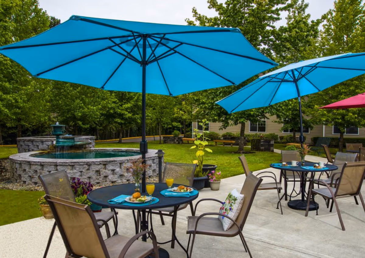 Outdoor patio with round tables and chairs under turquoise umbrellas next to a stone fountain and grassy lawn.