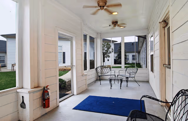 A screened-in porch area with white wooden walls and ceiling, two ceiling fans, black metal chairs and a small round table, a blue rug on the floor, and a fire extinguisher mounted on the wall near a glass door leading outside to a grassy courtyard with other buildings visible.