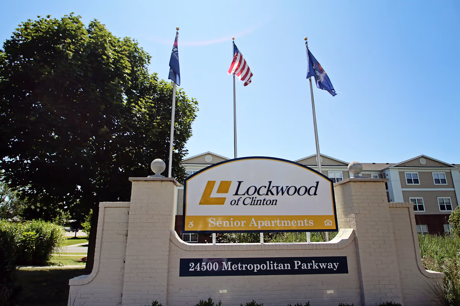 Outdoor view of the entrance sign for Lockwood of Clinton Senior Apartments located at 24500 Metropolitan Parkway, with three flagpoles flying flags behind the sign and a residential building in the background under a clear blue sky.