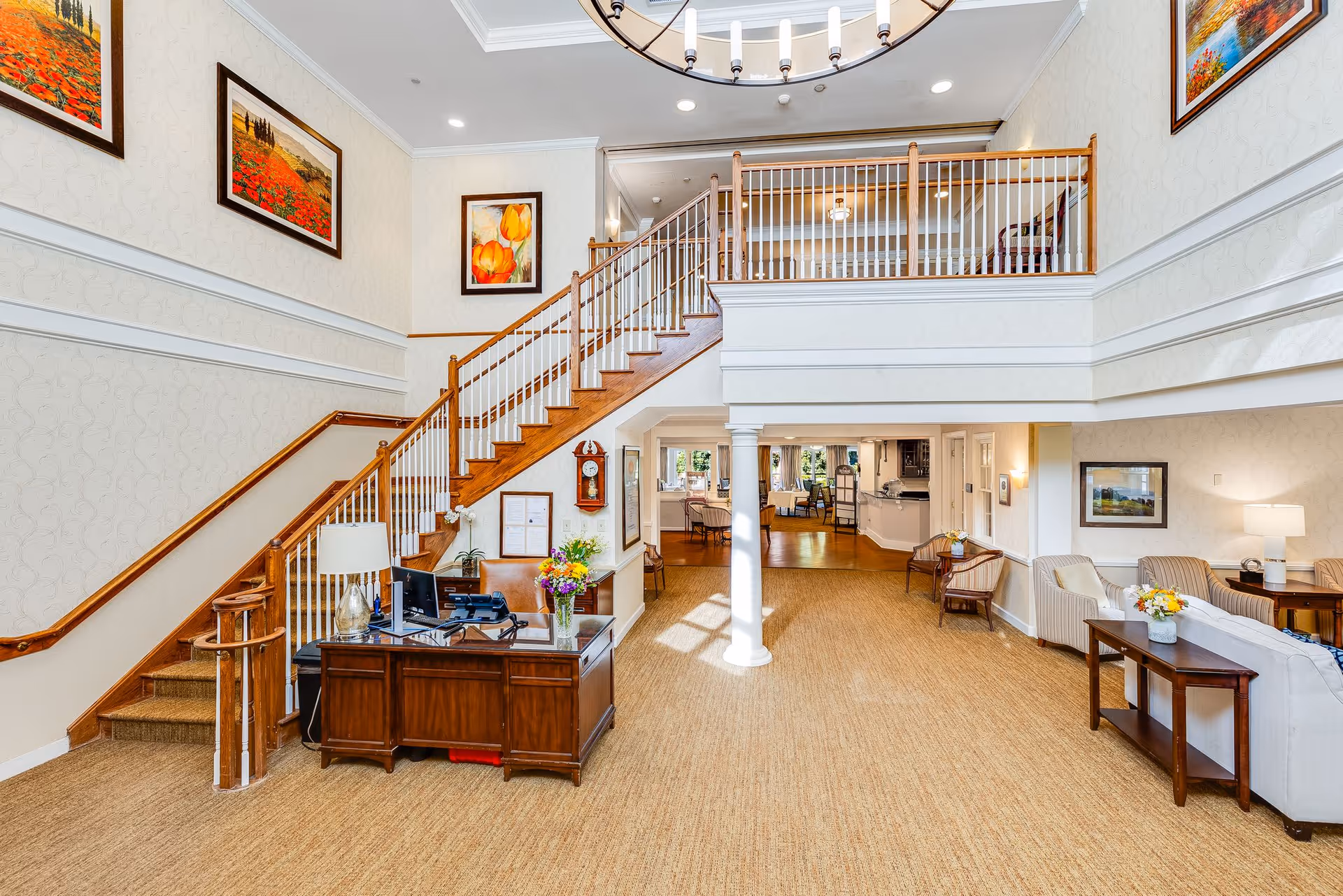 Bright and spacious senior living facility interior with a wooden staircase leading to an upper level. There is a wooden reception desk with a computer and a vase of flowers near the stairs. The room features beige carpet, white walls with decorative molding, framed floral and landscape paintings, and comfortable seating areas with chairs and sofas. A large chandelier hangs from the ceiling, and natural light fills the space.
