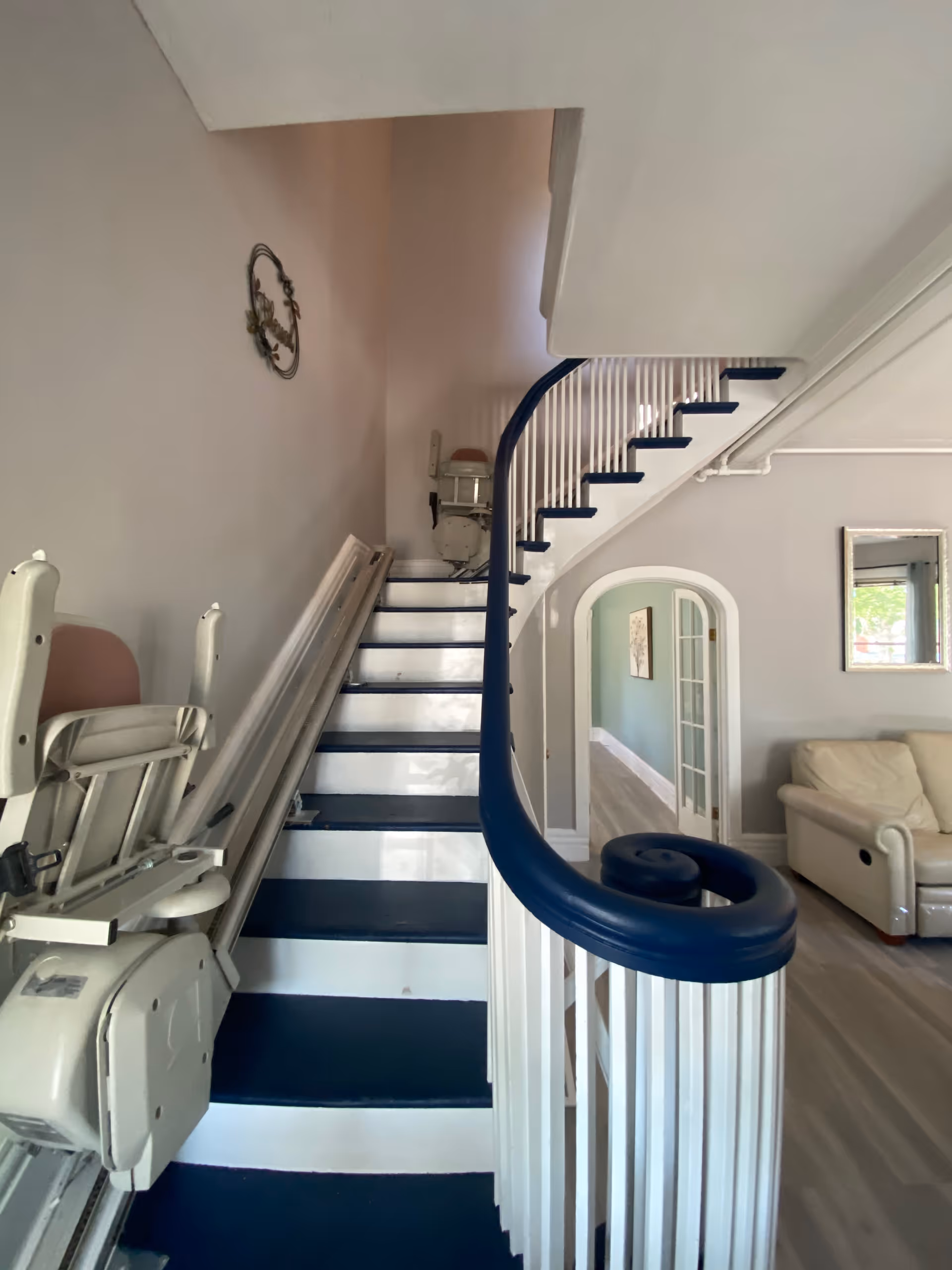 Curved indoor staircase with a blue handrail and a stairlift installed, leading past an archway into a living area.