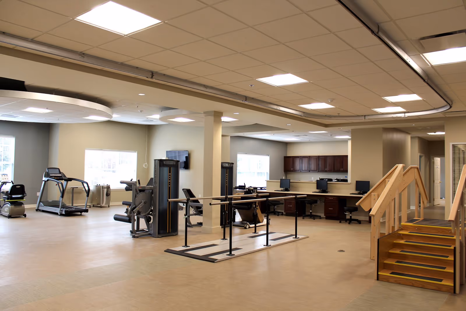 A spacious rehabilitation or physical therapy room with exercise equipment including treadmills, stationary bikes, and strength training machines. There are parallel bars for walking exercises and a set of wooden stairs for stair climbing practice. The room has large windows letting in natural light, a ceiling with recessed lighting, and a workstation area with computers and chairs in the background.