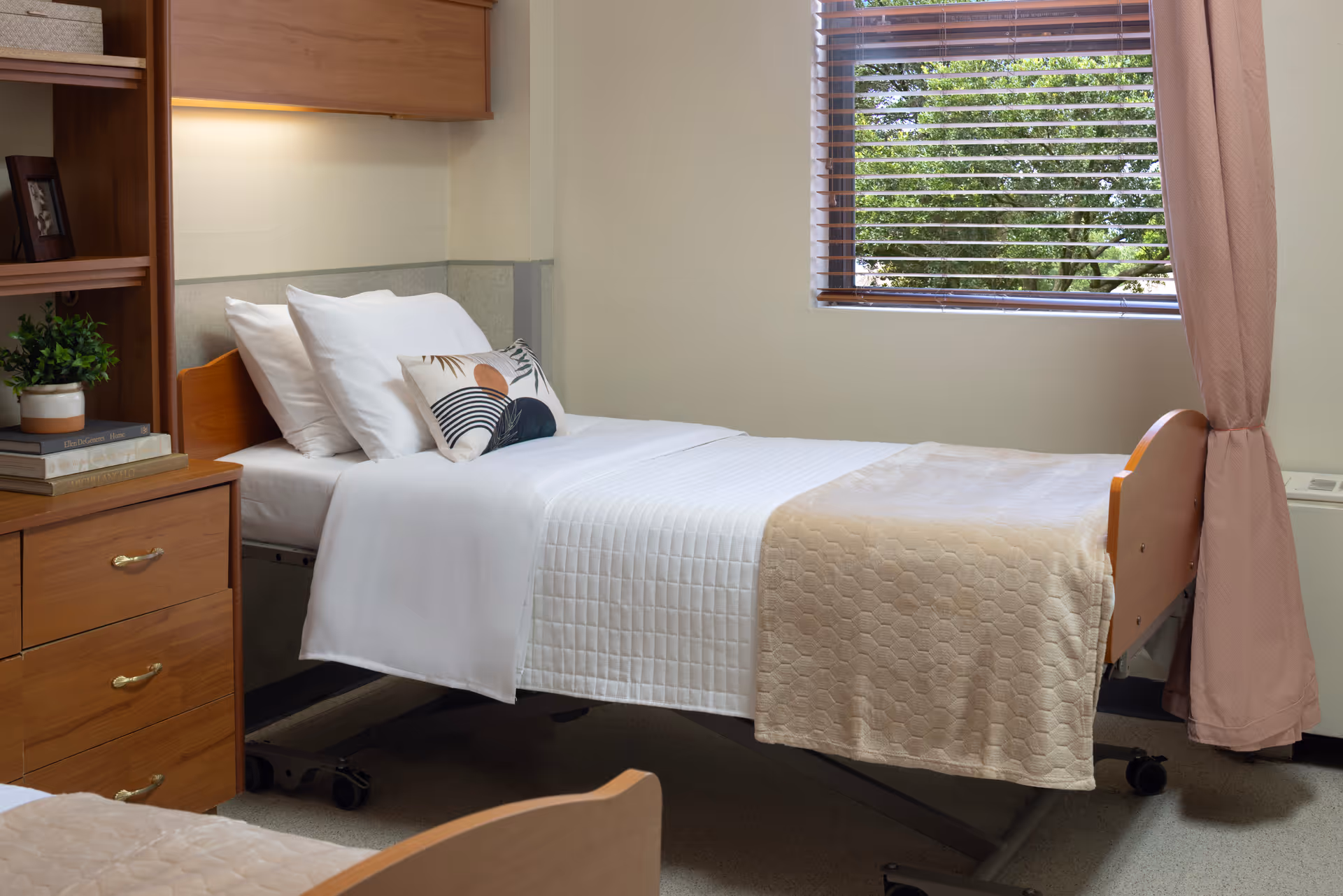 A neatly made single bed in a senior living facility room with white sheets, pillows, and a decorative cushion. The room features wooden furniture including a dresser and shelves with books and a small plant. A window with wooden blinds and pink curtains lets in natural light, showing green trees outside.